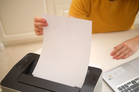 A woman with a laptop prints paper on a printer while sitting at a table in a home kitchen. An adult...