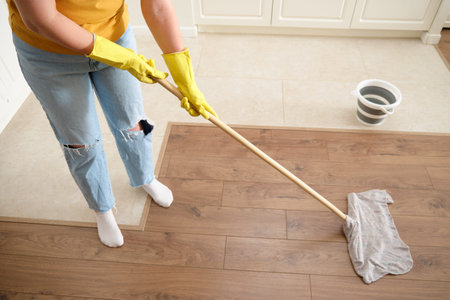 A woman mops the floor in her home kitchen. Female hands in yellow gloves while cleaning the kitchen