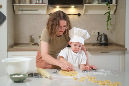 A woman and a child are making cookies together in a kitchen. The woman is wearing an apron and the...