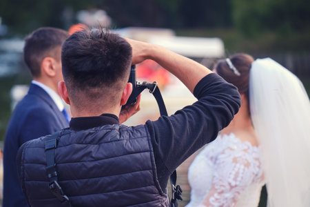 Photographer in black vest taking a photo of bride and groom during outdoor wedding shootの写真素材
