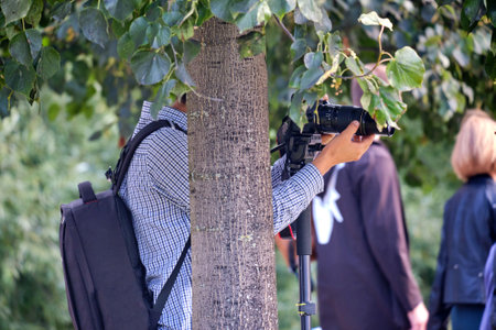 Photographer with camera on tripod partially hidden behind a tree in a park. Outdoor photography sessionの写真素材