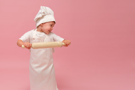 A young chef baby, wearing a hat and apron, smiles while holding a rolling pin in a portrait set against a pink background. Kid aged 3 years (three year old boy)の写真素材