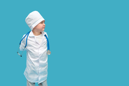 Baby doctor with a stethoscope on a studio blue background. A child chooses a medical profession by playing in a doctor's uniform. Kid aged three years (a three-year-old boy)の写真素材