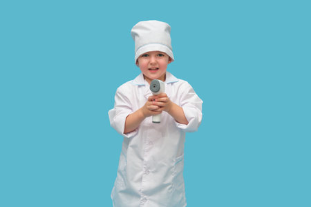 A baby doctor with a thermometer measures the temperature on a studio blue background. A child chooses a medical profession by playing in a doctor uniform. Kid aged three years (a three-year-old boy)の写真素材