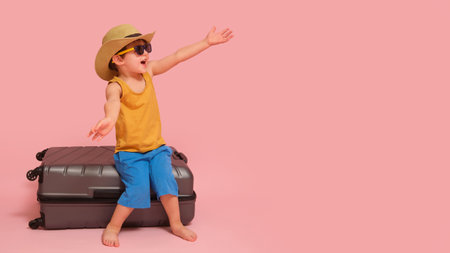 The baby boy happily rests on a suitcase packed with things for the tour, studio pink background. Kid aged three years (a three-year-old boy)の写真素材