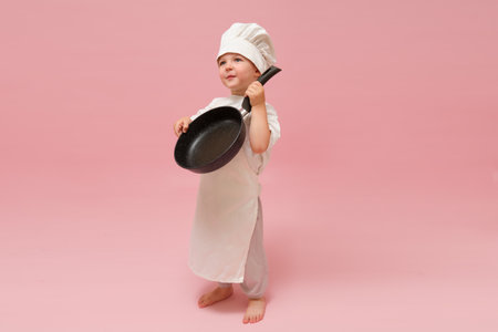 A happy baby wearing a chef's uniform is holding a cooking frying pan. Pink studio backdrop. Kid aged 3 years (three year old boy)の写真素材