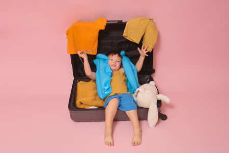 The baby boy is sitting in a suitcase, surrounded by clothes, eagerly waiting for a holiday trip, studio pink background. Kid aged three years (a three-year-old boy)の写真素材