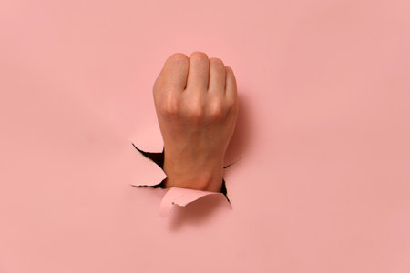 Hand breaking through pink paper wall with clenched fist. Conceptual studio shot for creative design.の写真素材
