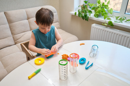 The child is molding plasticine with his hands at the table in the cozy home living room. A toddler baby is playing with colorful playdough, crafting imaginative shapes indoors. Kid aged three yearsの写真素材