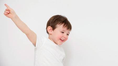 Smiling child pointing upwards with raised arm on white background. Studio portrait photography with copy space. Childhood happiness and playful moment concept. Design poster, banner, greeting cardの写真素材