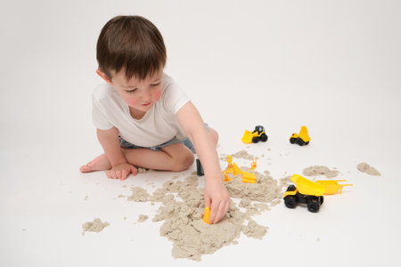A happy child is playing indoors with kinetic sand, building cars and bulldozers. The toddler baby is engaged in a fun learning experience with kinetic sand on a white studio. Kid is three years oldの写真素材