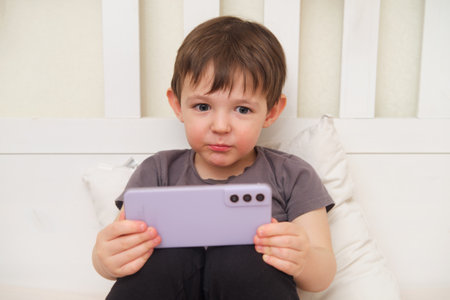 A young child lying on a pillow and holding a phone in the bedroom. An adorable baby holding a smartphone while resting on a pillow in bed. Kid aged three (three-year-old)の写真素材
