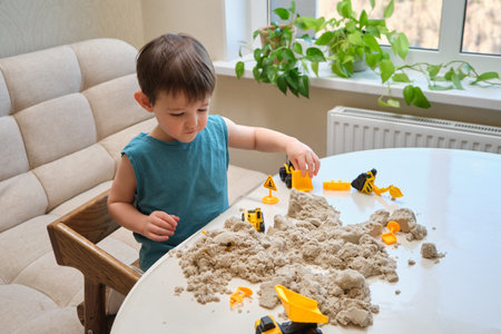 Child playing with kinetic sand and toy construction vehicles on a white table. Indoor activity in a bright room with green plants by the window. Childhood creativity and sensory play concept.の写真素材