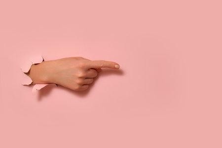 Female hand pointing through torn pink paper on a pink background. Studio photography with copy space. Creative direction and gesture concept for design and print.の写真素材