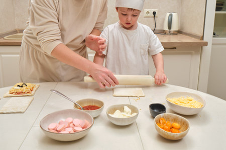 A woman and her child are rolling out pizza dough, sharing a fun cooking experience. The mom, acting like a chef, helps her little boy prepare pizza with lots of flour on the table.の写真素材