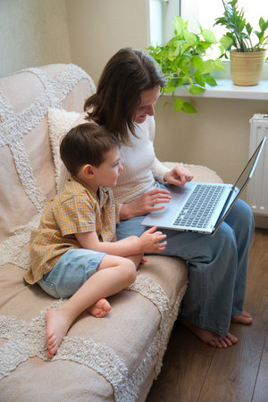 Mother and son sitting on a cozy sofa using a laptop at home. Family bonding and online learning concept.の写真素材