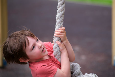 Child holding onto a rope on a playground with a blurred background. Happy kid training on a climbing rope at a park. A child aged 3 yearsの写真素材