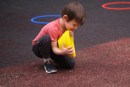 Young child squatting on playground, holding yellow cones with a joyful expression, surrounded by colored hoops on rubber surface. A child aged 3 yearsの写真素材