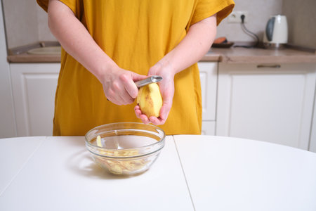 Woman peeling raw potato with metal peeler over transparent glass bowl containing potato peels. Studio kitchen scene. Home cooking and meal preparation concept. Close-up viewの写真素材