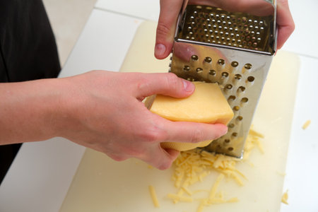 Close-up of hands grating a block of hard cheese using a stainless steel box grater on a kitchen counter.の写真素材