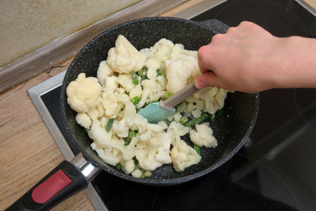 A person stirs chopped cauliflower and green beans in a non-stick frying pan on an electric stove, preparing a healthy meal.の写真素材