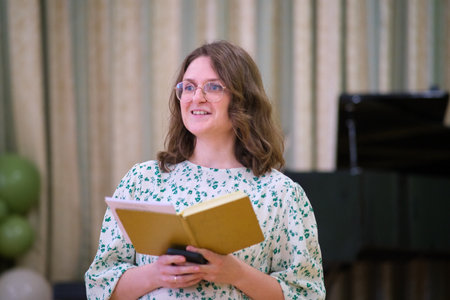 Smiling woman with glasses holding a yellow notebook and smartphone, speaking or reading during an indoor public or formal event.の写真素材
