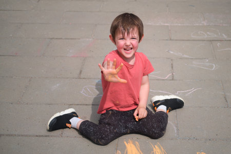 Happy child sitting on concrete floor, extending hand in playful gesture while drawing with sidewalk chalk. Outdoor play and childhood fun concept. A child aged 3 yearsの写真素材