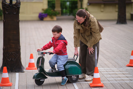 Woman helping a boy to ride a toy scooter. Kid learns to balance a scooter on a wooden path. Childhood education and parenting. A child aged 3 yearsの写真素材