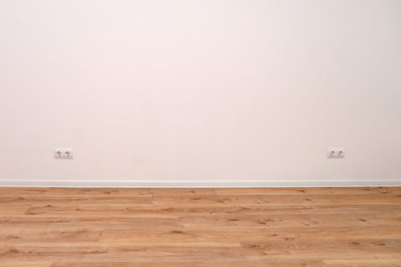 Empty room with white wall, wooden floor, and two electrical outlets. New home interior with natural...