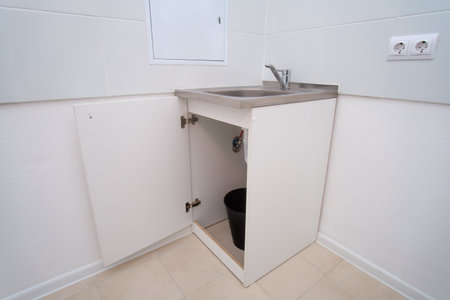 White laundry room or utility room with an open cabinet under a stainless steel sink, revealing a black bucket inside.の写真素材