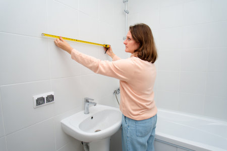 A woman measures the width of a tiled bathroom wall using a yellow tape measure, preparing for renovation or home improvement project.の写真素材