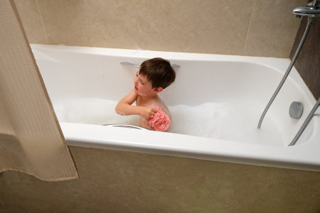 Little boy child enjoying bubble bath with pink sponge for personal hygiene and daily routine during...