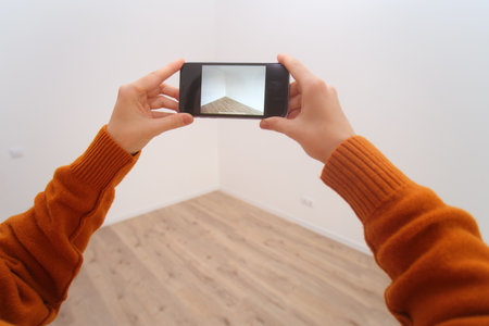Woman holding smartphone to capture photo of empty, renovated room with light wood floor and white walls. Moving or real estate concept.の写真素材