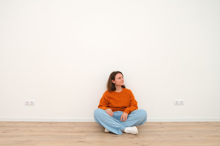 Woman sitting cross legged on wooden floor, looking away with smile. Caucasian female in empty room with white wall, copy space.の写真素材
