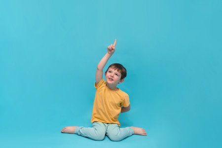 Little kid pointing up with an index finger, sitting on a blue background. Boy looking towards copy space. Child advertising or promoting.の写真素材