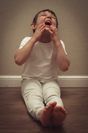 Little boy crying out loud while sitting on floor against wall.の写真素材