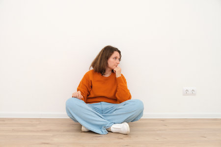 Thoughtful woman sitting on floor with legs crossed. Caucasian female student contemplating on problem concept. Minimalist room with copy space.の写真素材