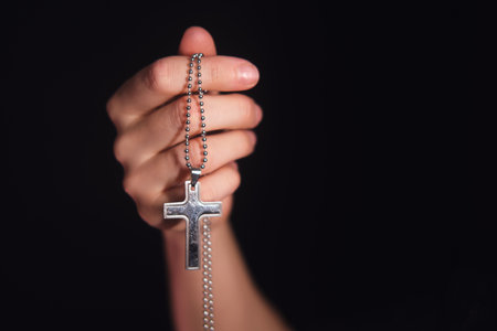 Hand of caucasian child holding silver cross on dark background. Christian religious symbol for faith, prayer, and spiritual concept with copy space.の写真素材