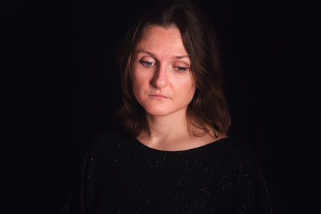 Woman looking down with a sad or pensive expression. Portrait of an adult female with brown hair on a black background for emotional concept.の写真素材