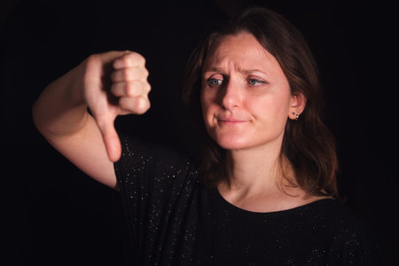 Young woman making a thumbs down gesture with a dissatisfied facial expression on a dark background. Concept of disapproval for negative feedback or rejection.の写真素材