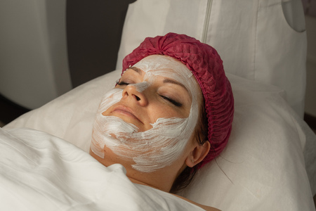 The cosmetologist for procedure of cleansing and moisturizing the skin, applying a sheet mask to the face of a young woman in beauty salon. Cosmetology and professional skin care.の写真素材