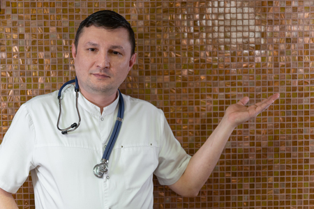 Doctor advertising your product. Cheerful mature doctor looking at camera and pointing away while standing against gold backgroundの写真素材