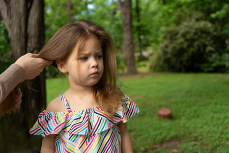 Mother's hands puts her daughter's thick hair, takes care of her hair. Woman brushing her daughter's hair outdoorsの写真素材
