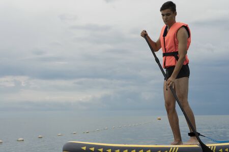 Young man in a life jacket goes surfing. The concept of water safety and learning to surf with a paddleの写真素材