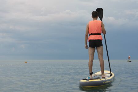 Young man on sup board in a life jacket on the background of the sea. View from back surferの写真素材