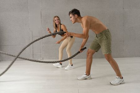The personal trainer is engaged with the client on a battle rope in gym. Athletic female doing some cross-training exercises with her clientの写真素材