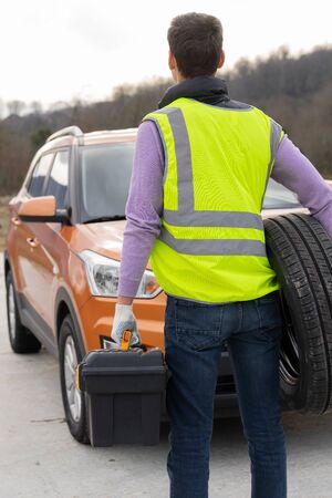 Photo of a driver with tools to repair the car and a spare wheel in his hands, in front of his vehicle. The man is dressed in high-visibility or reflective vestの写真素材