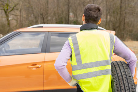 The driver in a reflective vest with a spare wheel on the background of the car