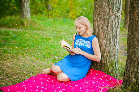 blonde girl in a blue dress sitting on a red blanket on top of the green grass, reading a book in the middle of pine forest treesの写真素材