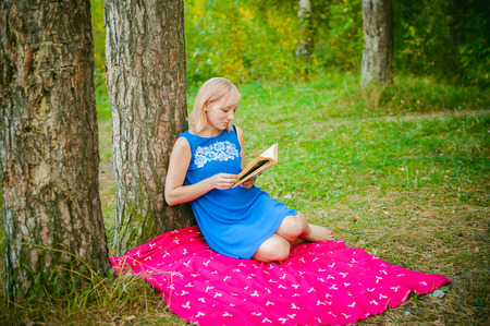blonde girl in a blue dress sitting on a red blanket on top of the green grass, reading a book in the middle of pine forest treesの写真素材